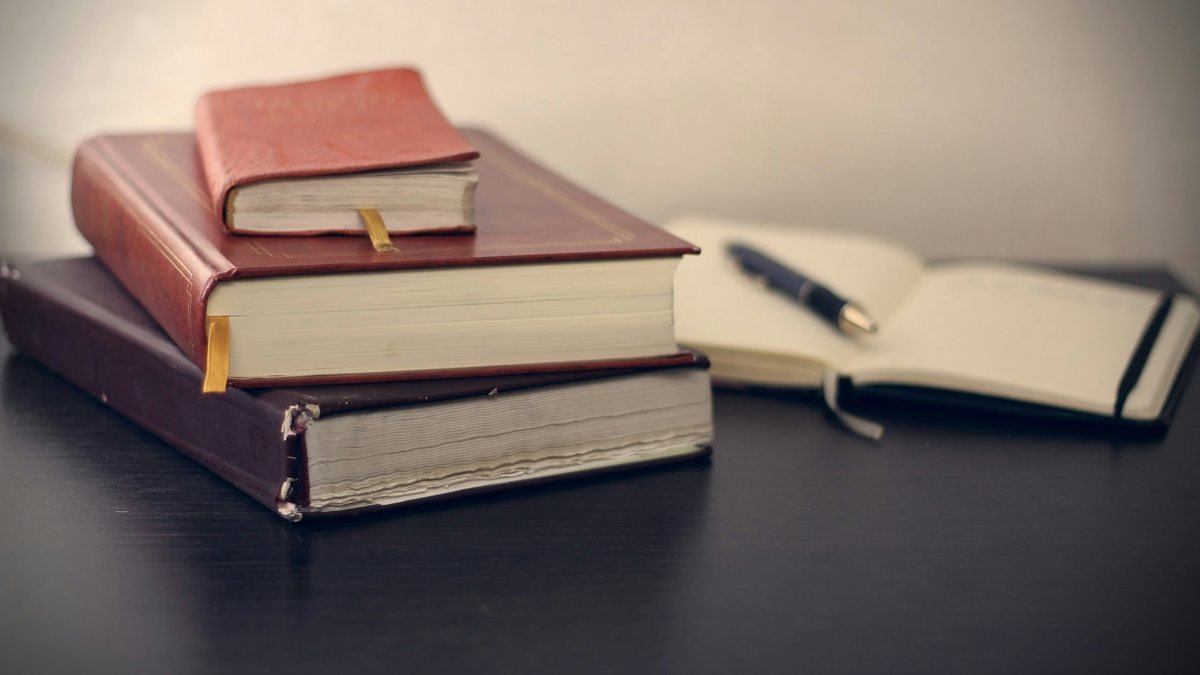 Livros empilhados ao lado de um caderno aberto com uma caneta, sobre uma mesa escura.