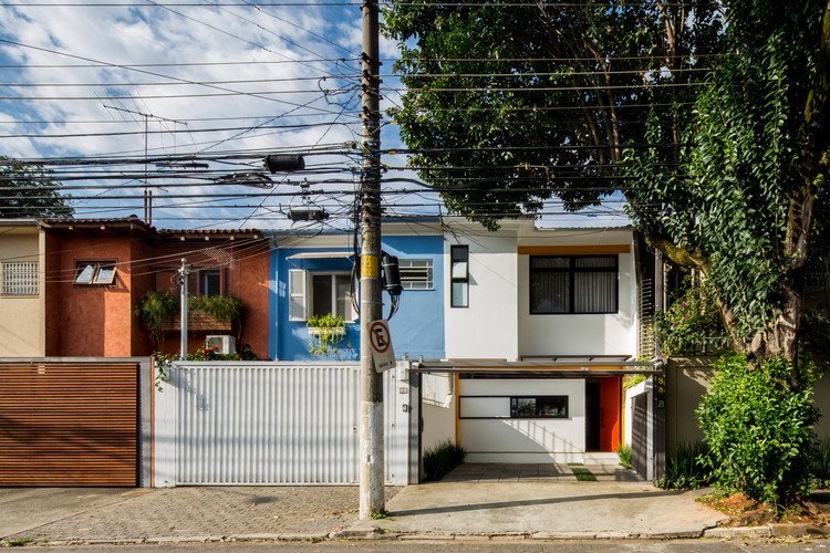 Rua de bairro com casas e pequenos comércios de fachada colorida, com fios de poste e vegetação.