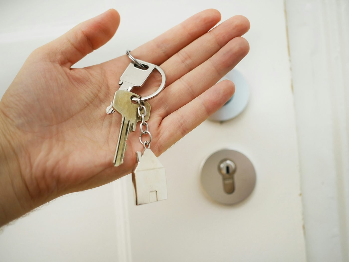 Mão segurando um molho de chaves com chaveiro em formato de casa, diante de uma porta.